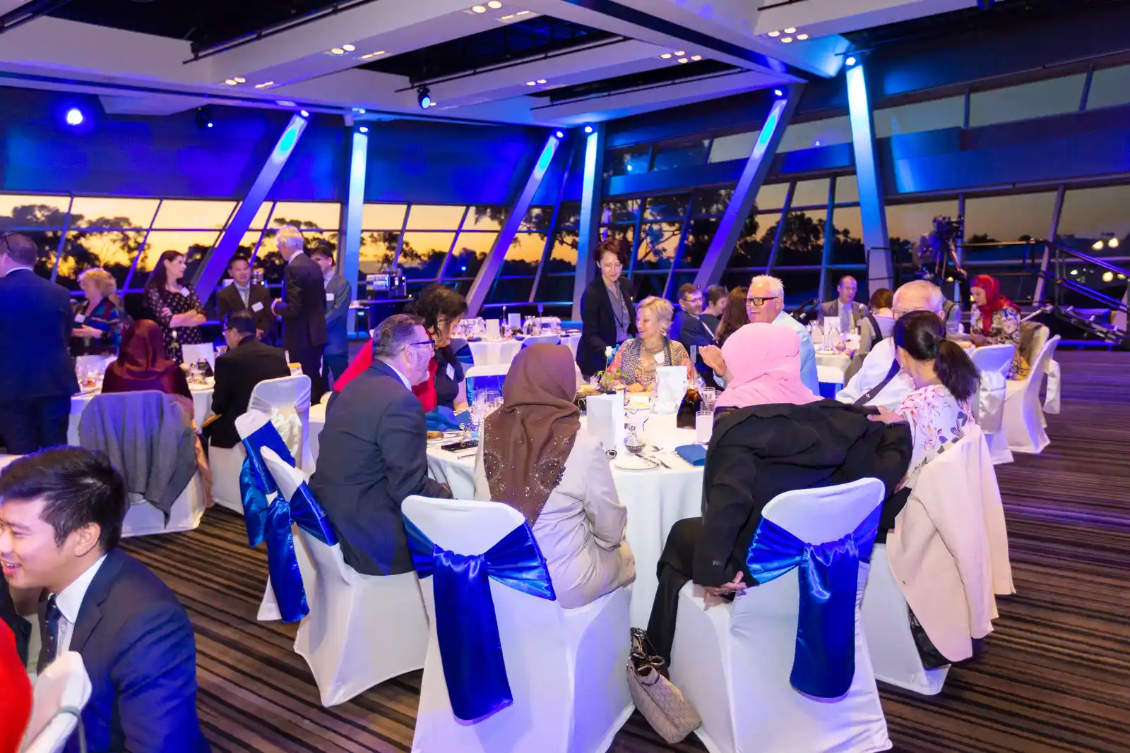 Gala dinner guests at round tables in a function room with floor-to-ceiling windows at sunset