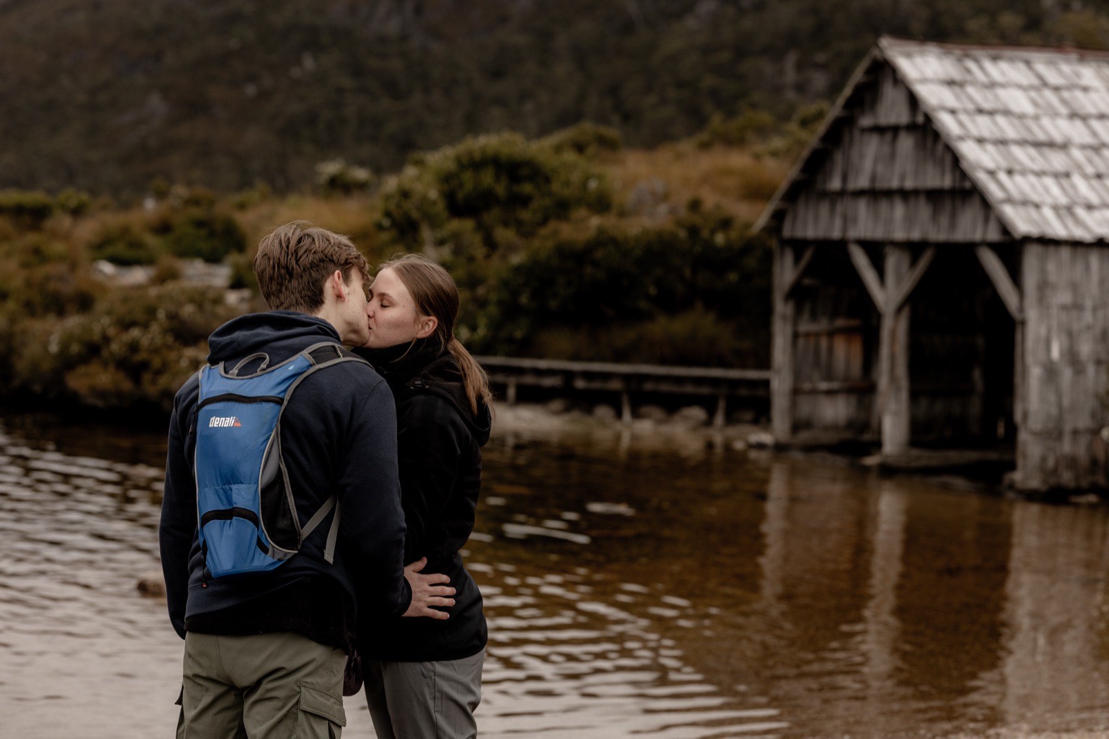 Newly engaged couple kissing by the water with the Dove Lake boatshed behind them
