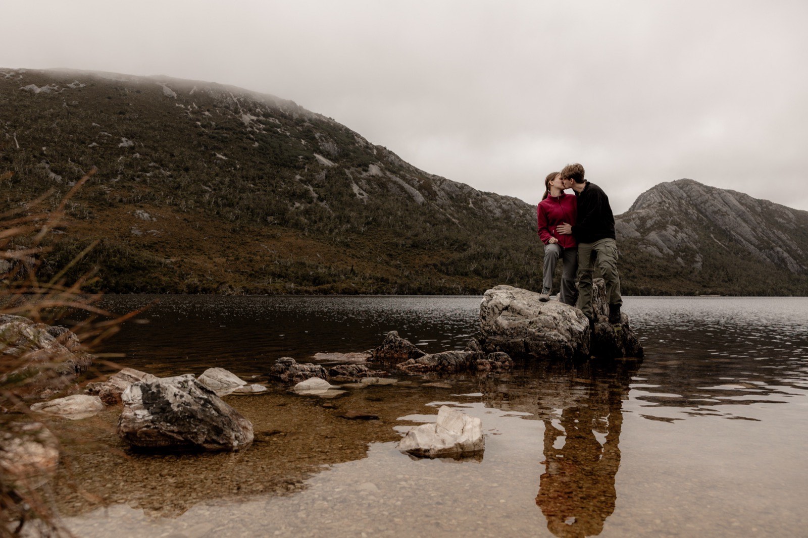 Engaged couple standing on a rock in Dove Lake, kissing, with Cradle Mountain reflected in the water